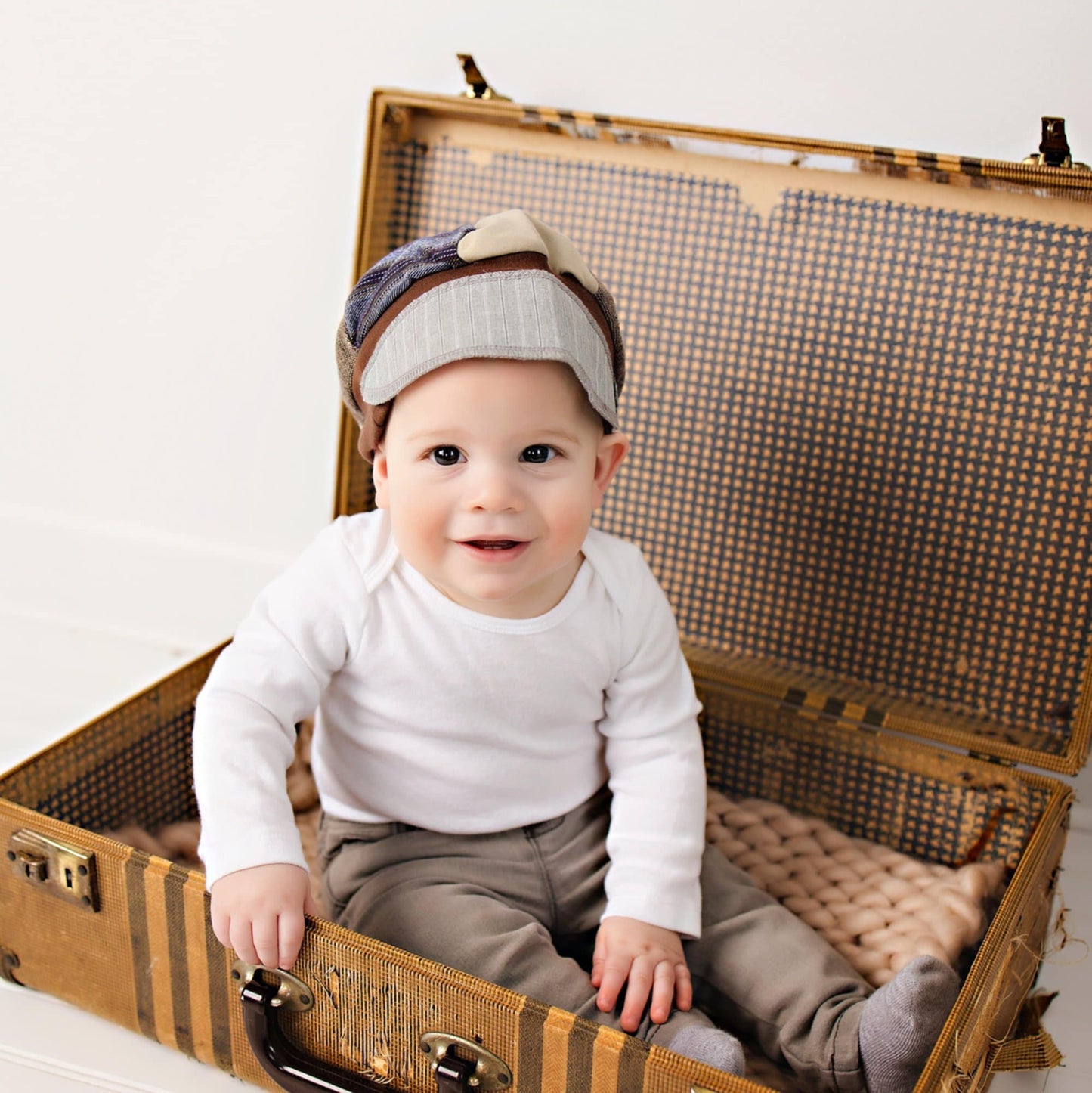 Baby sitting in an open vintage suitcase wearing a white shirt and gray pants with a checkered hat.