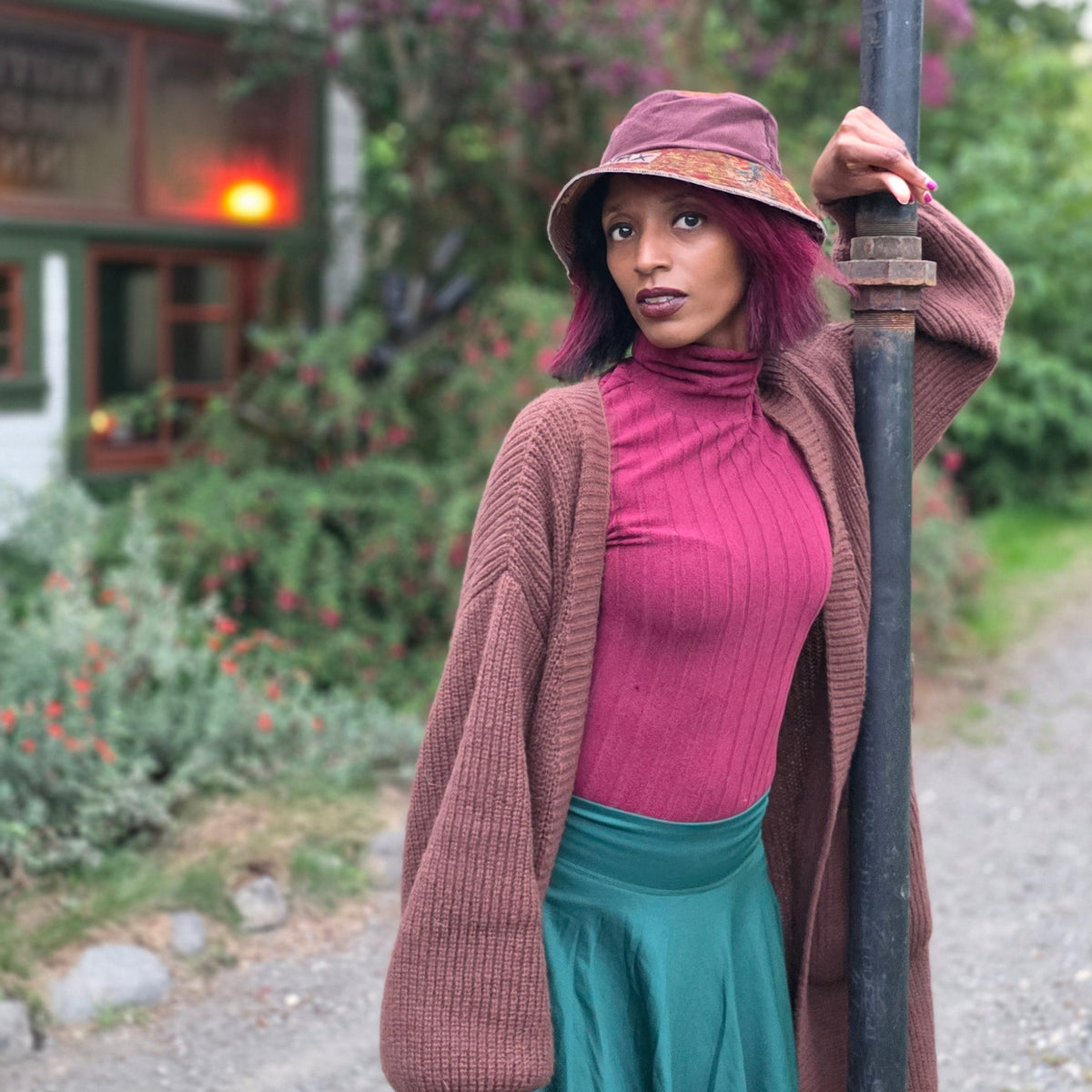 A woman posing outdoors in a rustic setting, wearing a brown knit cardigan, a magenta ribbed turtleneck, a teal mini skirt, and a brown bucket hat while leaning against a black metal pole.