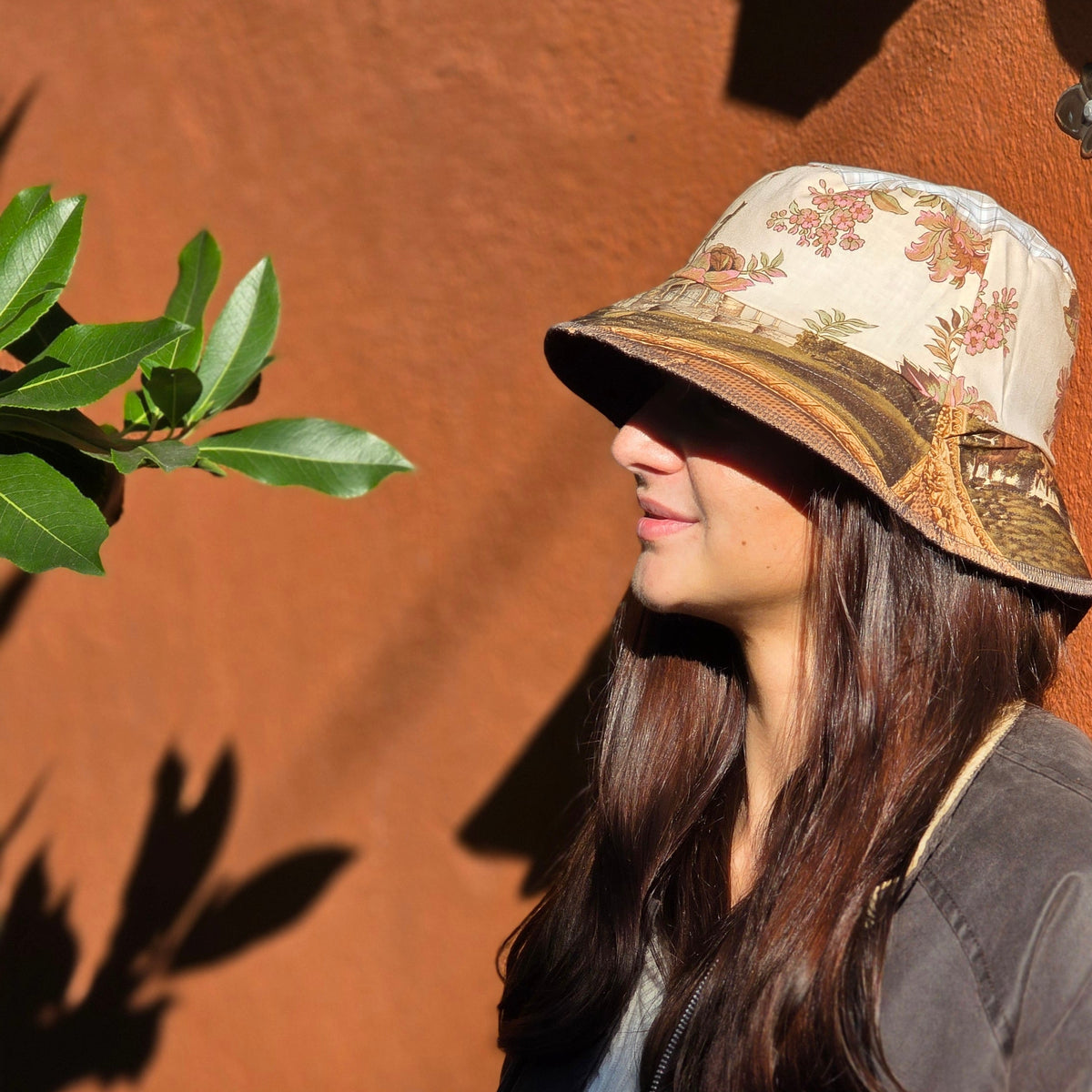 A woman wearing a beige bucket hat with a brown floral pattern and a dark brown jacket, standing against a textured terracotta wall with leaf shadows.