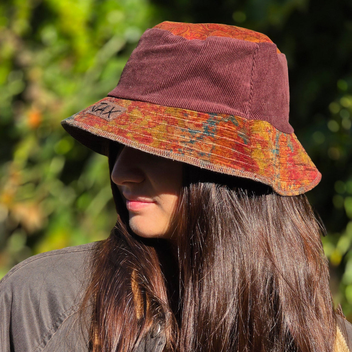 A woman wearing a wide-brimmed bucket hat with a maroon crown and a rust-orange patterned brim, photographed outdoors against a blurred green background.