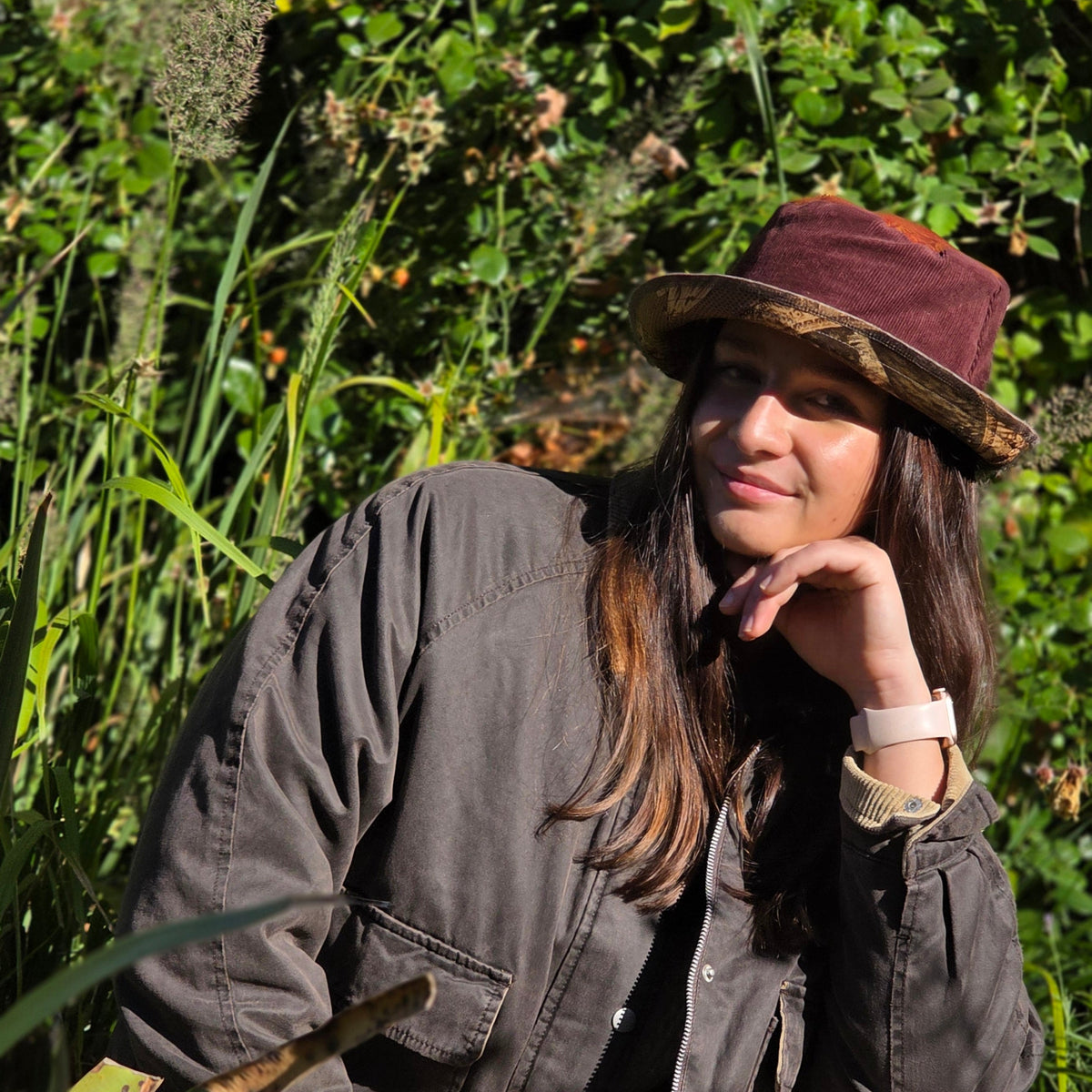 A woman outdoors in a lush green field, wearing a dark gray utility jacket, a maroon corduroy hat with a camouflage brim, and a light pink smartwatch.