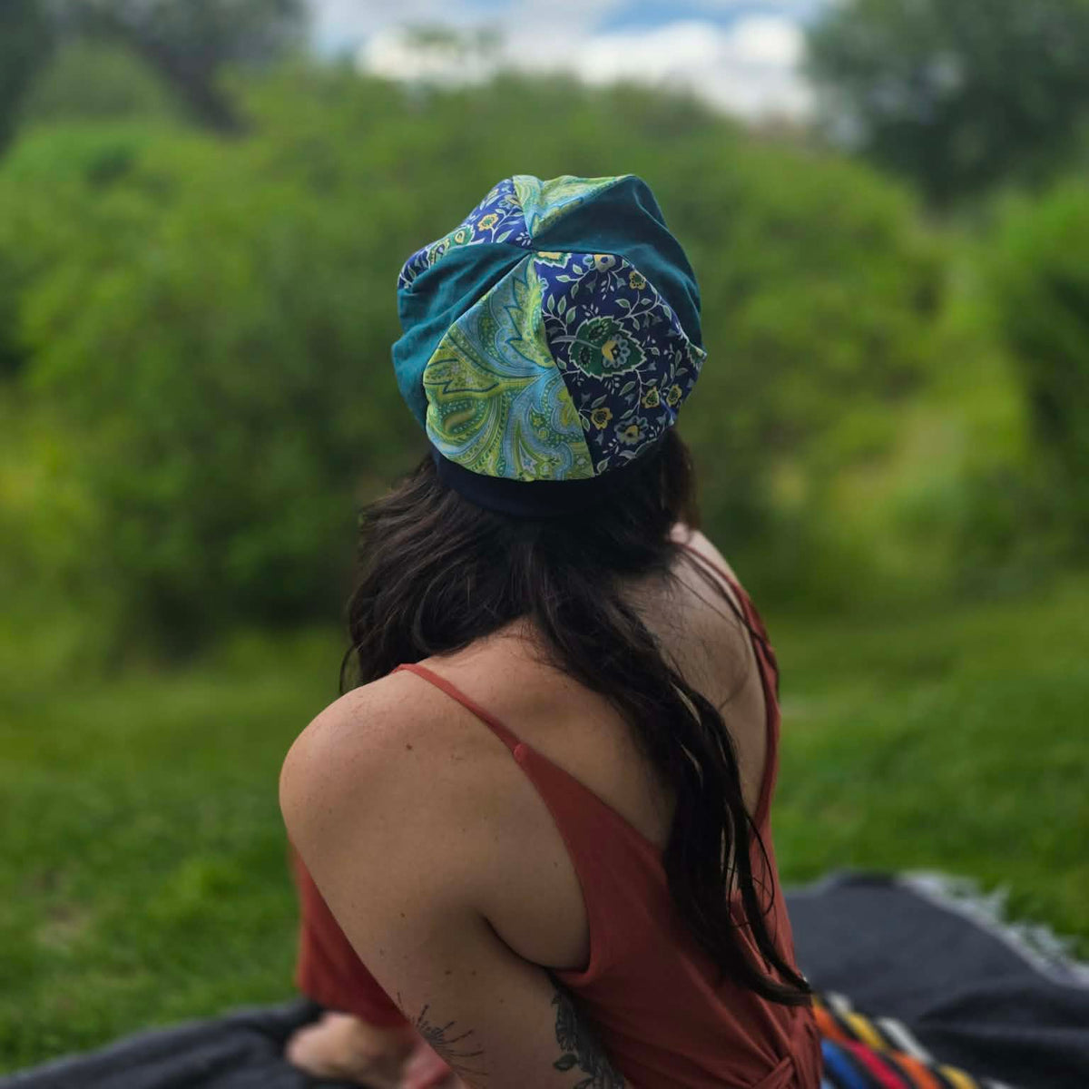 A woman seen from behind wearing a colorful patterned fabric headwrap outdoors in a grassy park with trees and a cloudy sky in the background.