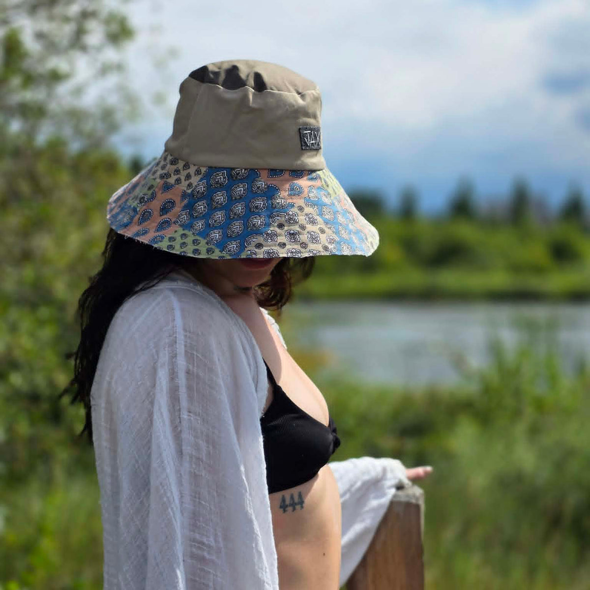 A woman wearing a beige wide-brim sun hat with a colorful floral patterned brim, a white open-front shirt, and a black bra standing outdoors near a wooden railing with a lake and greenery in the background.