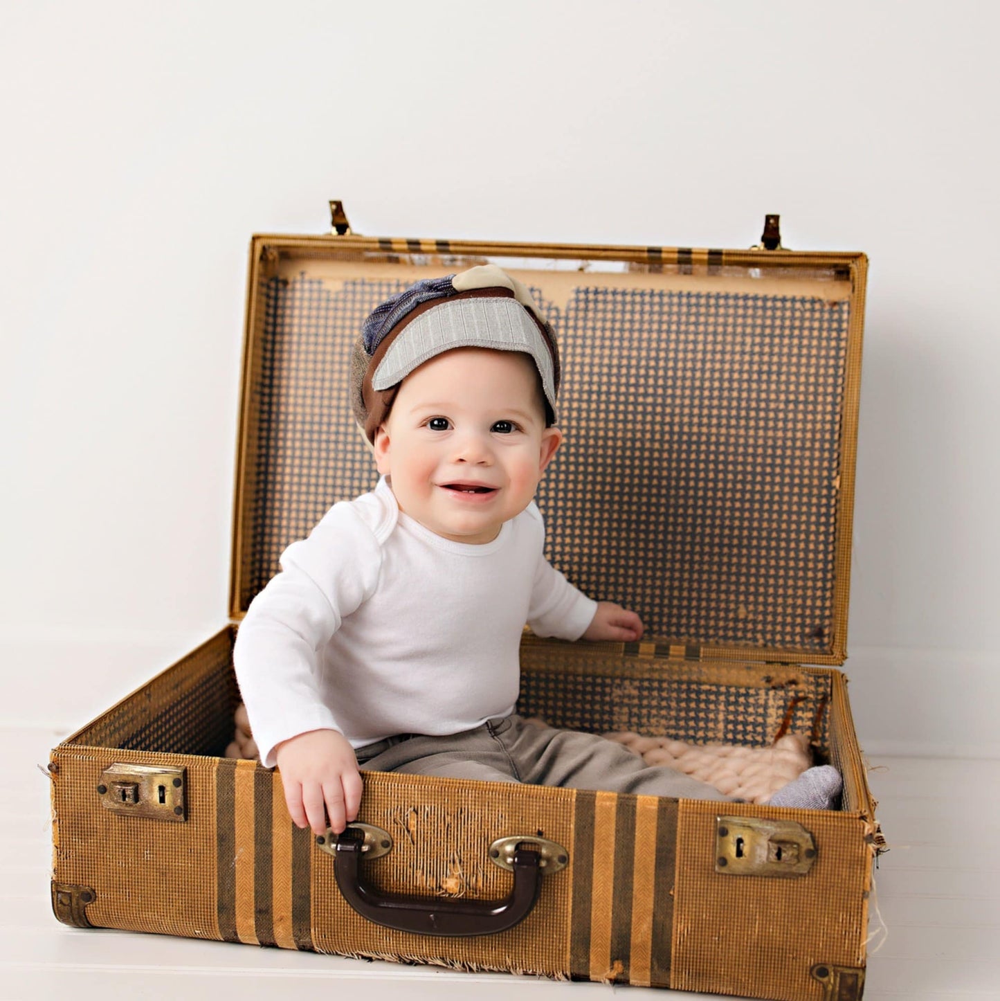 Baby sitting inside an open vintage suitcase on a plain background wearing a patchwork upcycled newsboy cap