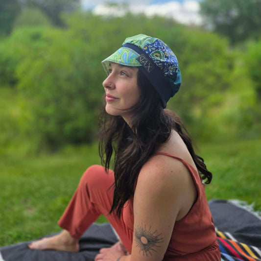 A woman sitting outdoors on a blanket in a grassy field, wearing a colorful patterned sun hat with blue, green, and yellow tones, along with a rust-colored sleeveless top and pants.
