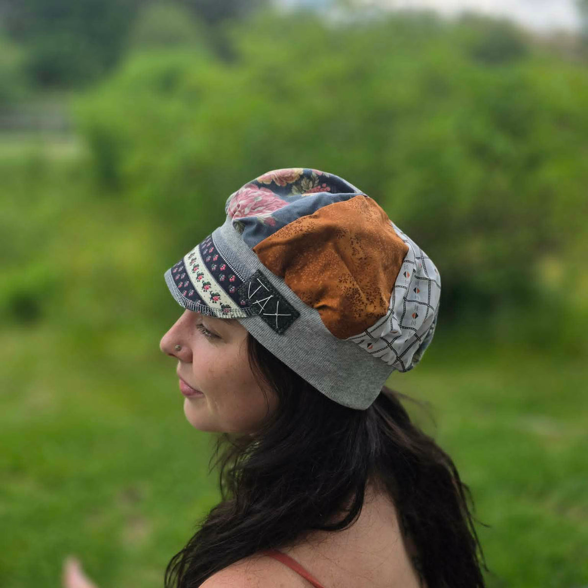 A woman in profile wearing a handmade patchwork beanie hat outdoors in a grassy park setting. The beanie features various fabric panels including a rust-orange textured section, a gray knit section, a patterned blue section, and a black-and-white geometric band.