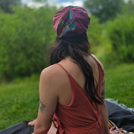 A woman sitting outdoors on a blanket in a grassy park, viewed from behind. She is wearing a rust-colored sleeveless dress and a maroon headwrap with colorful abstract patterns.