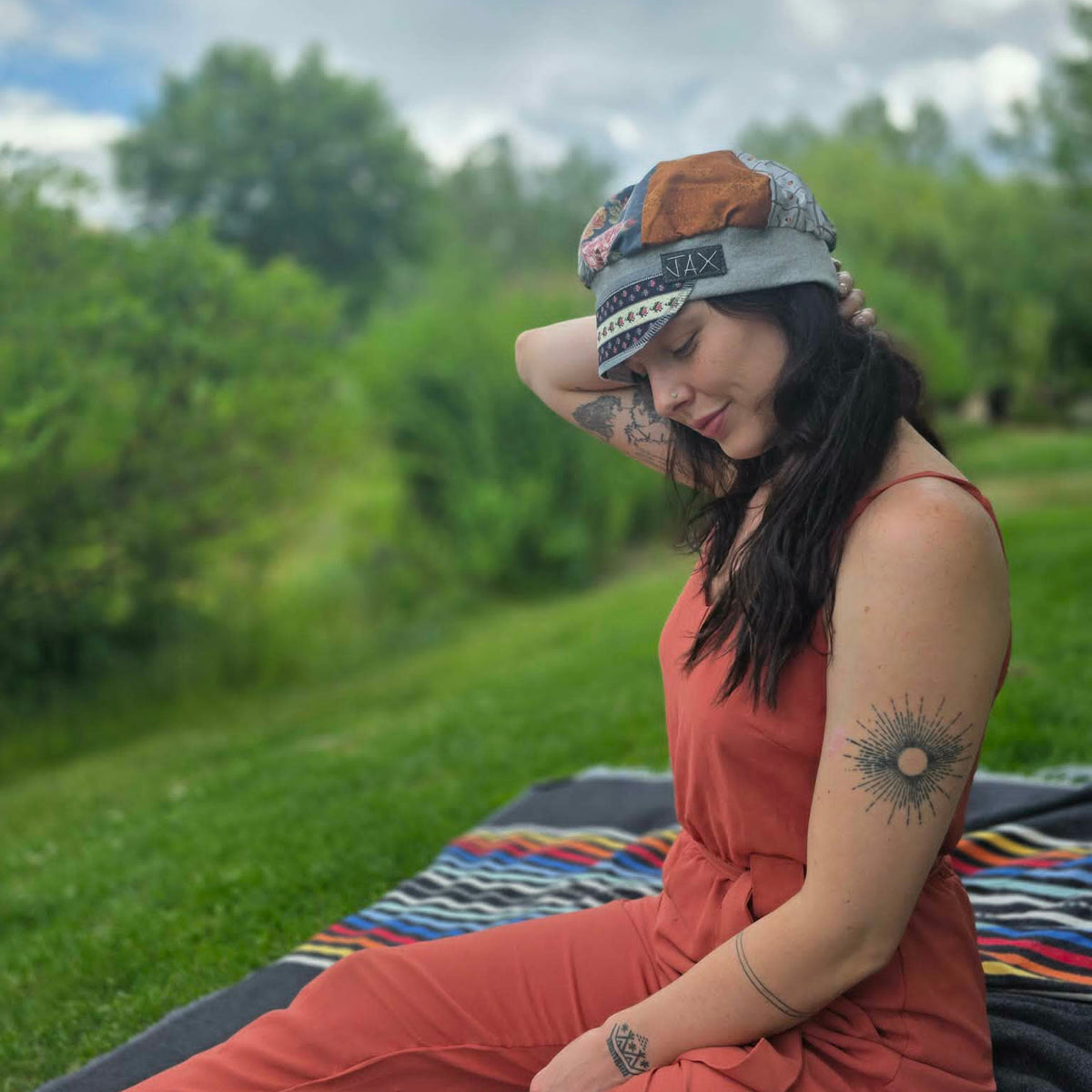 A woman sitting outdoors on a blanket in a grassy park setting, wearing a rust-orange sleeveless jumpsuit and a multicolor patchwork cap.