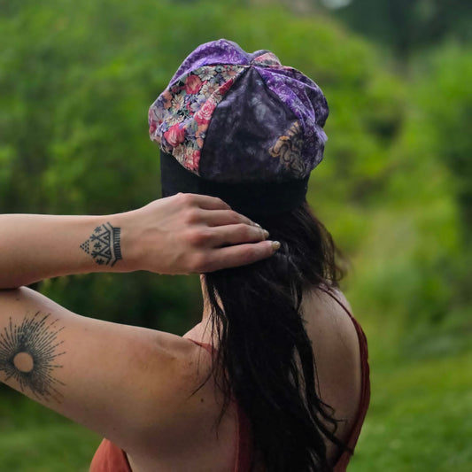 A woman viewed from behind outdoors, wearing a purple and floral patterned beret-style hat while adjusting her hair. The background is a soft-focus green natural setting.