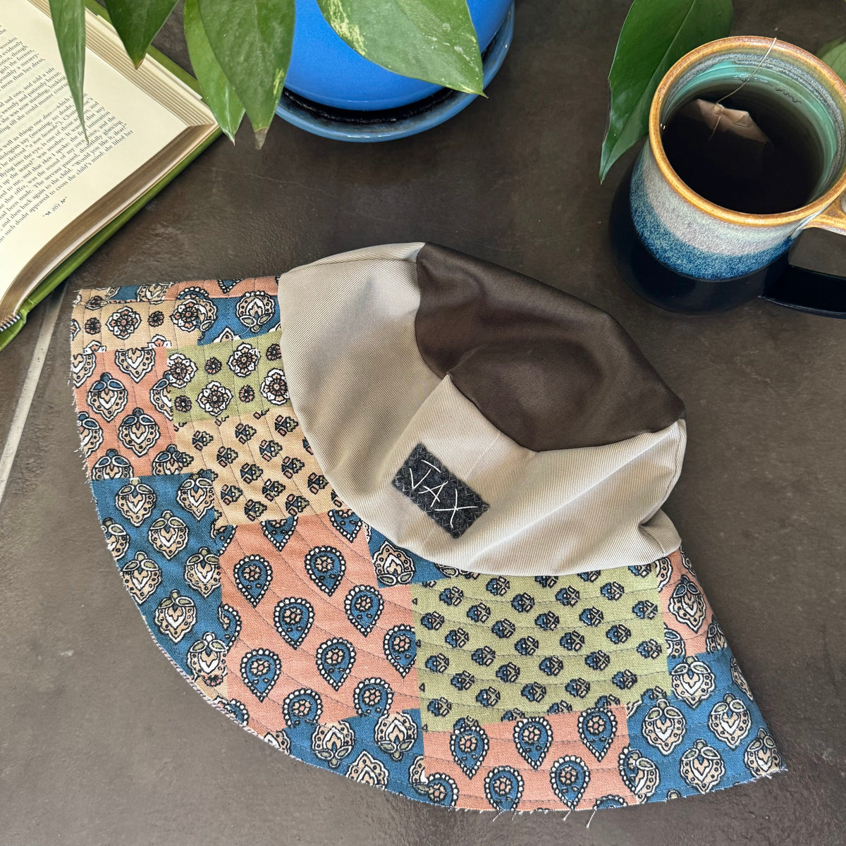 A handmade patchwork sun hat with a curved brim, featuring multiple colorful patterned panels (blue, pink, green, beige) and a solid beige crown, styled on a gray tabletop with a mug, book, and potted plants.