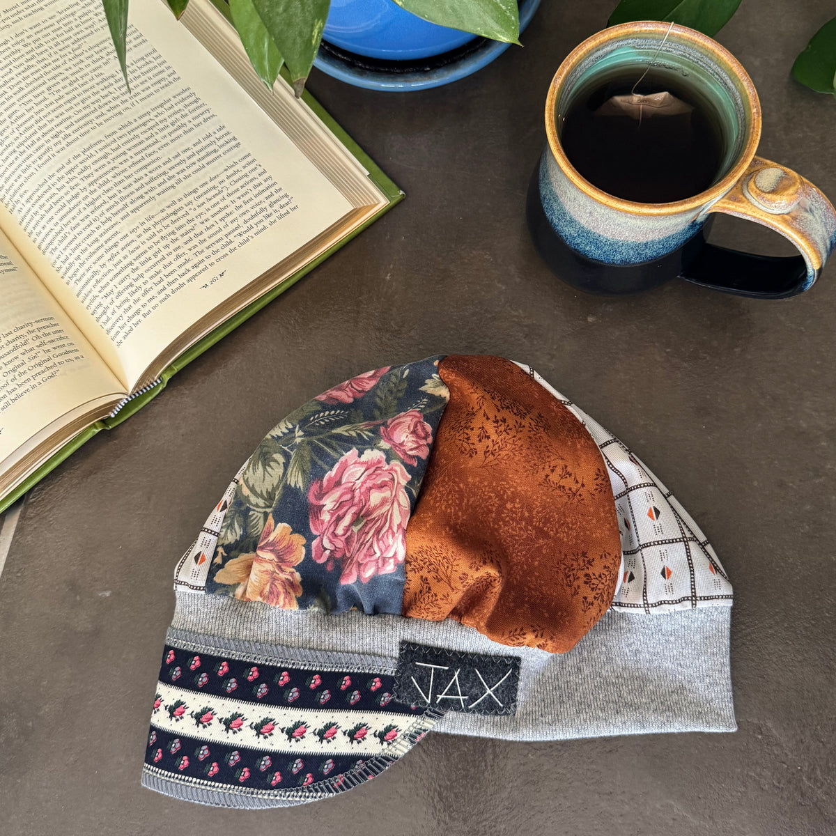 A folded knit beanie with mixed fabric panels (floral print, rust-brown textured fabric, and gray checkered fabric) displayed on a gray tabletop next to an open book, a mug of coffee, and a potted plant.