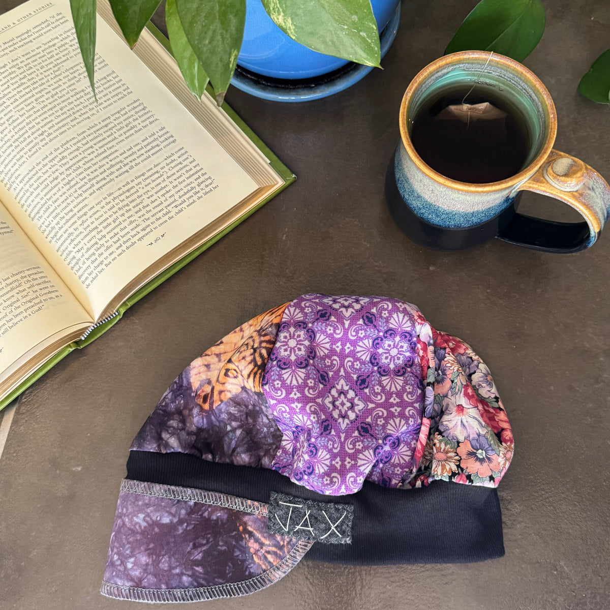 A patterned fabric hat or cap with purple, orange, and black designs, displayed on a gray tabletop next to an open book, a blue potted plant, and a ceramic mug of coffee.