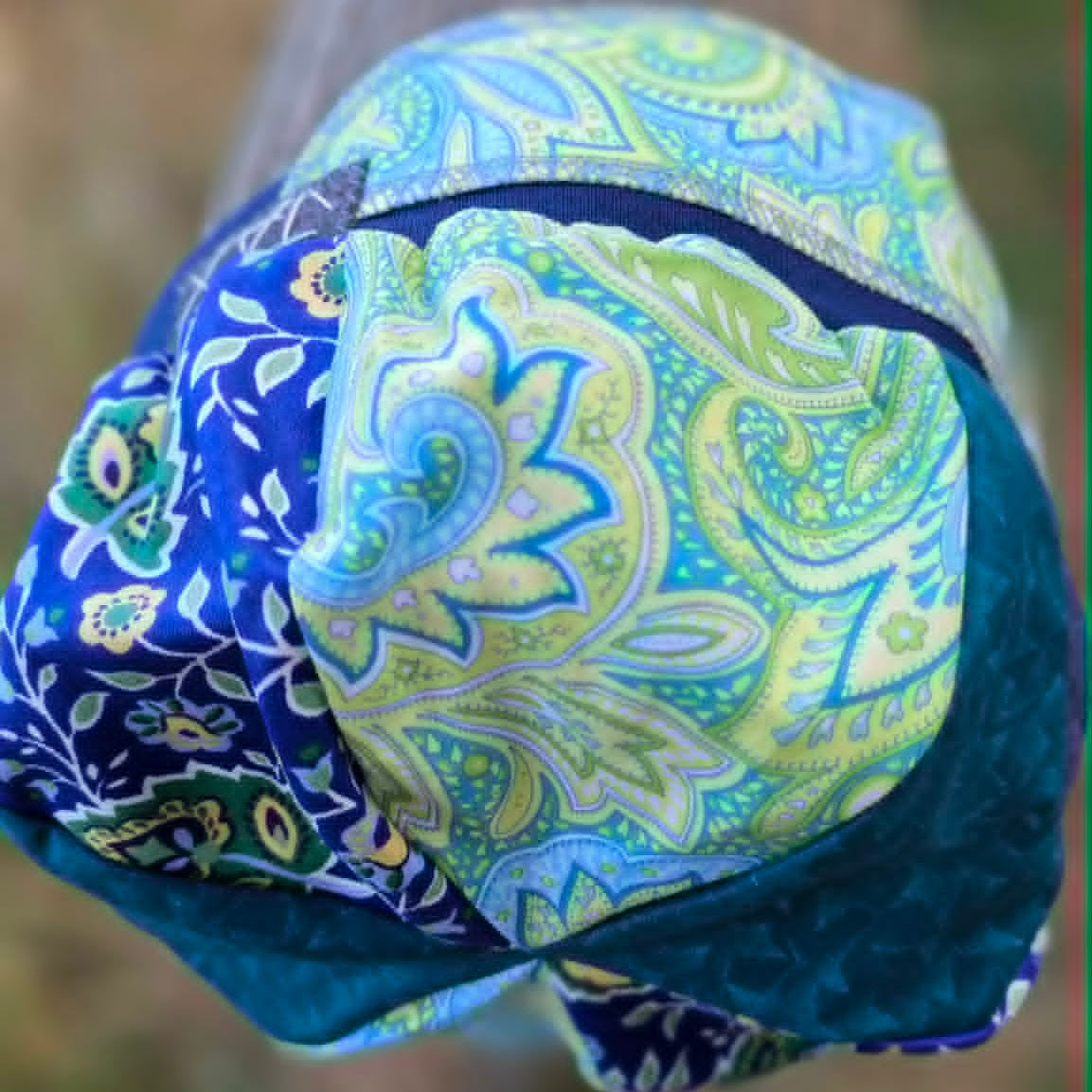 A close-up of a folded fabric headwrap or turban with a blue and green paisley pattern, draped over a rustic wooden post outdoors.