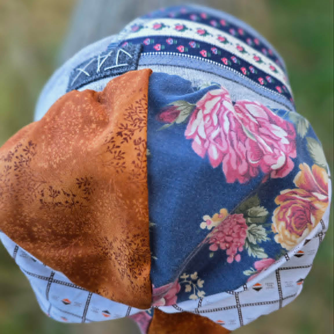 A close-up of a handmade patchwork fabric hat, likely a bonnet or small cap, featuring a mix of blue floral, brown suede-like fabric, and white checkered trim, photographed outdoors against a blurred background.