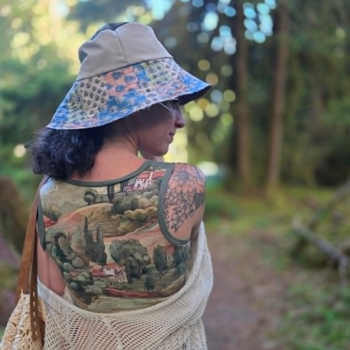 A woman outdoors on a forest path, shown from the back wearing a beige wide-brim sun hat with a blue patterned brim, a large landscape tattoo across her upper back, and a cream crochet wrap.