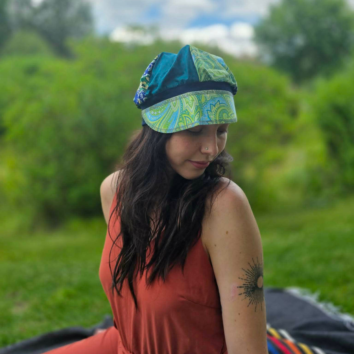 A woman wearing a teal and green paisley patterned sun hat and a rust-orange sleeveless dress, sitting outdoors in a grassy field under a cloudy sky.