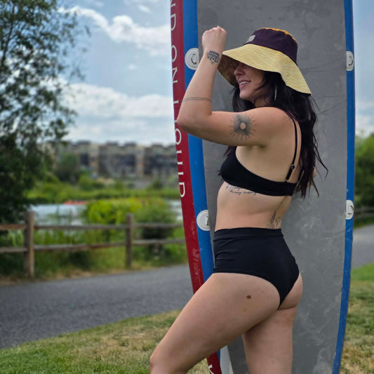 A woman in a black bikini and a yellow sun hat poses outdoors next to a surfboard, with grass, trees, and a cloudy sky in the background.