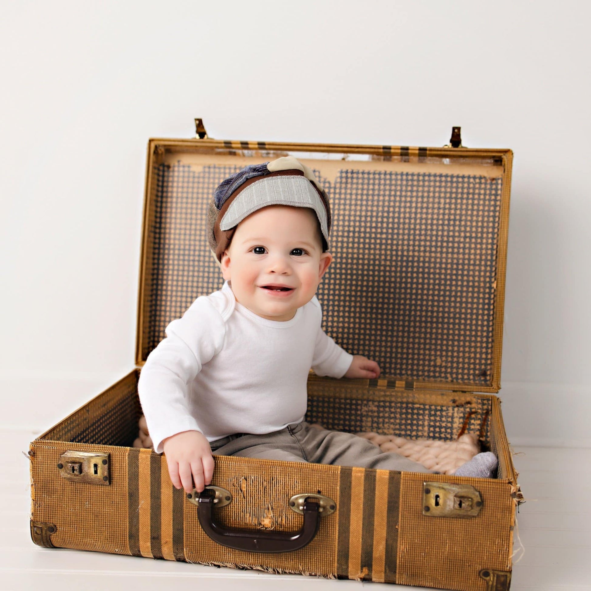 Baby sitting inside an open vintage suitcase on a plain background wearing a patchwork upcycled newsboy cap