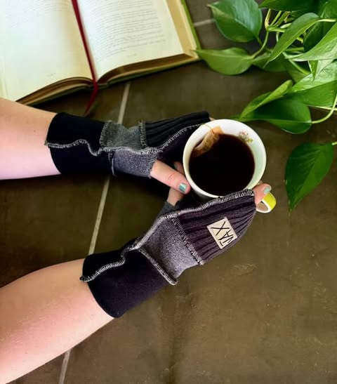 Person wearing black fingerless gloves holding a cup of coffee with a book and plant in the background.