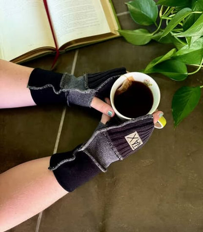 Person wearing black fingerless gloves holding a cup of coffee with a book and plant in the background.