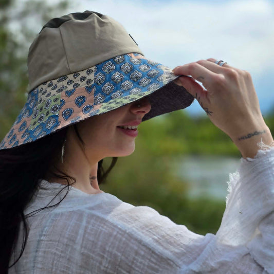 A woman wearing a beige wide-brim sun hat with a blue, pink, and white patterned brim, photographed outdoors against a blurred background of greenery and sky.